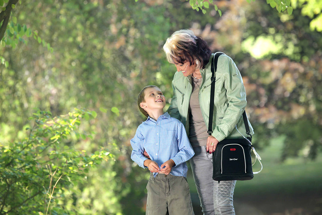 Old woman wearing nasal cannula connected to a GCE Zen-O Lite Portable Oxygen Concentrator and talking to a young boy